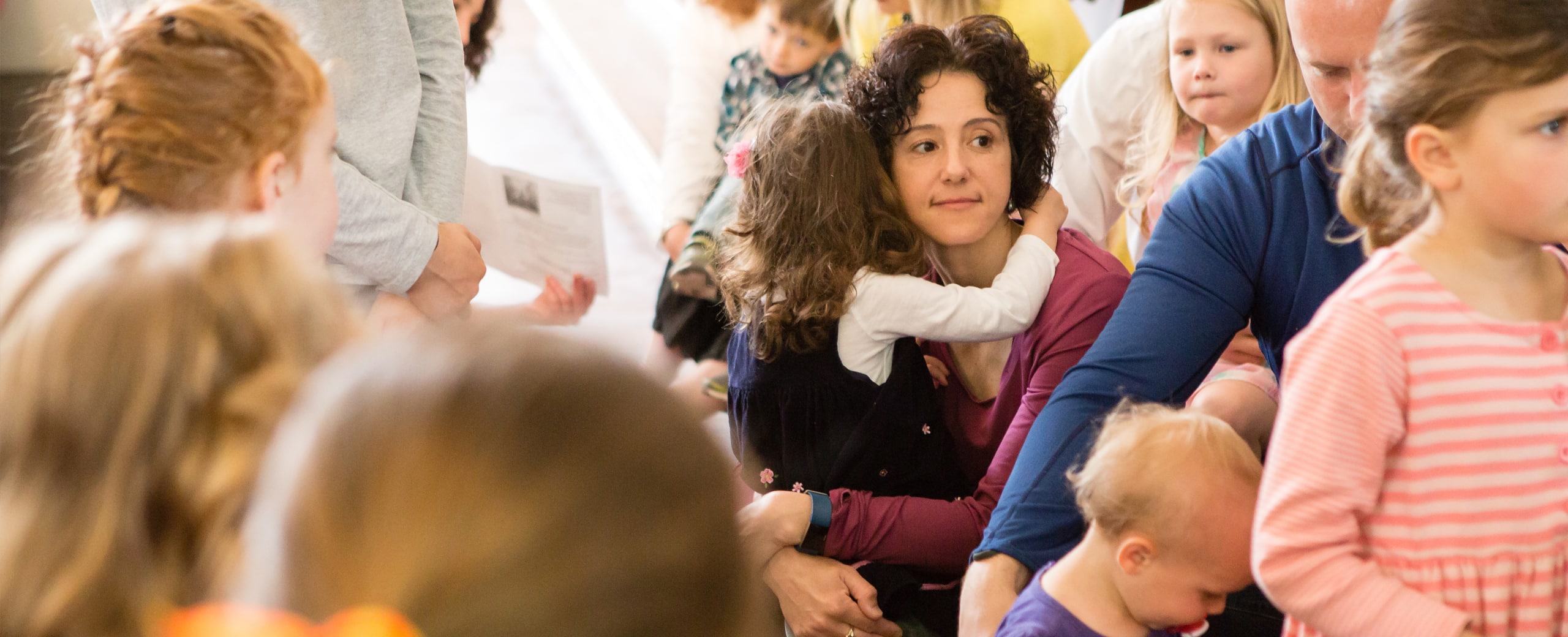 A mother hugs her daughter during the children's sermon. Other parents and children surround them.