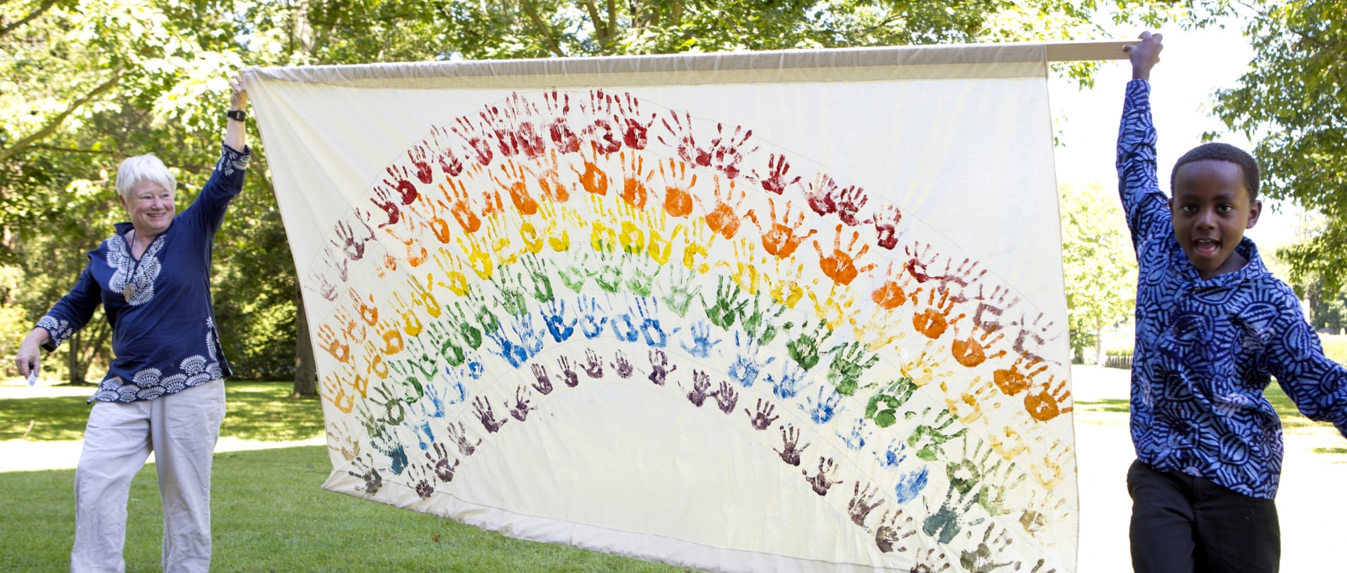 A middle-aged woman and a young African American boy hold up a rainbow banner made with children's hand prints.