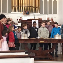Rev. Patty Fox, children, and their parents gathered around the communion table