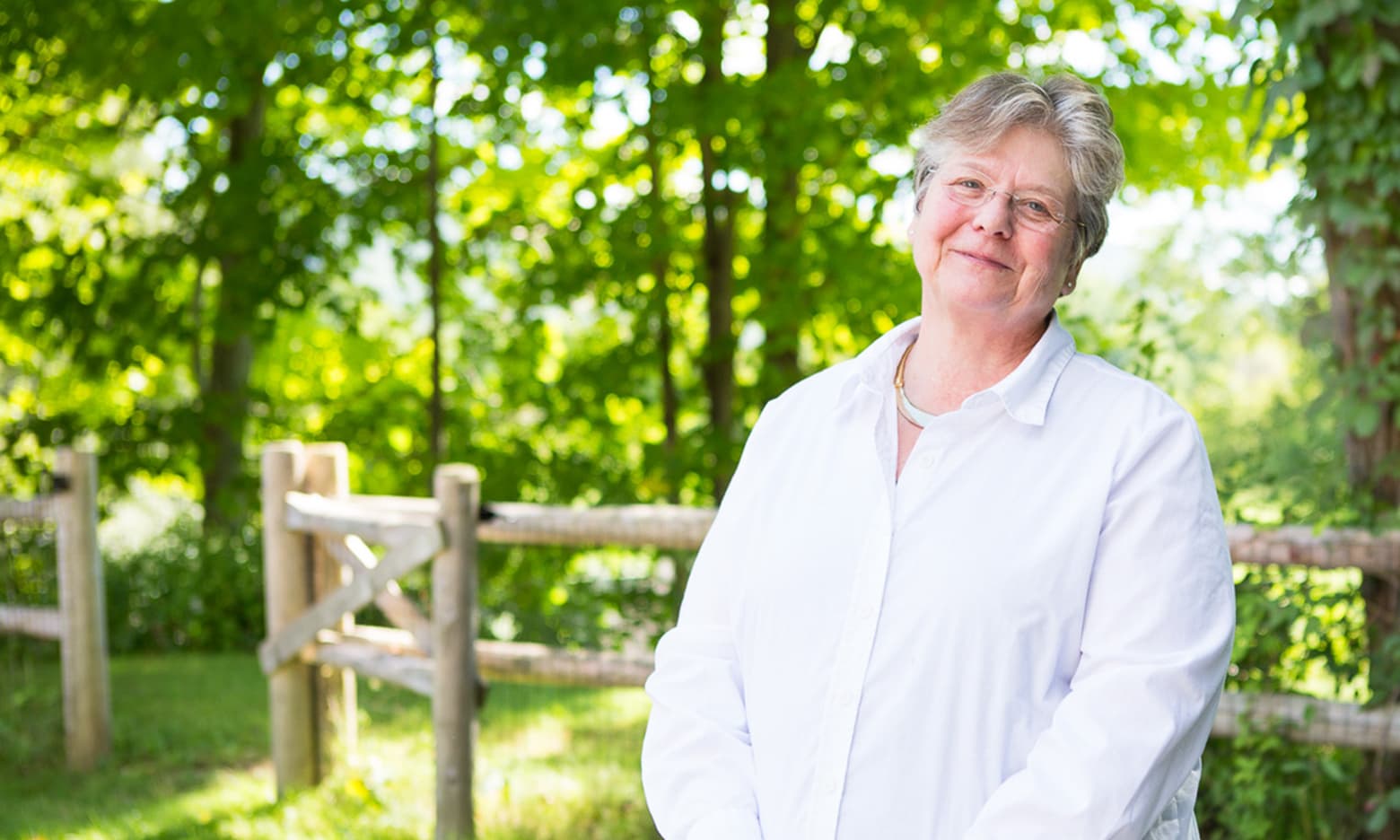A woman clasps her hands and smiles with green trees in the background