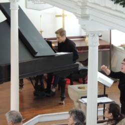 A young man plays the piano while the choir director conducts