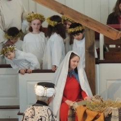 The wise men kneel before the manger. Mary adores the baby Jesus while children dressed as angels look on.